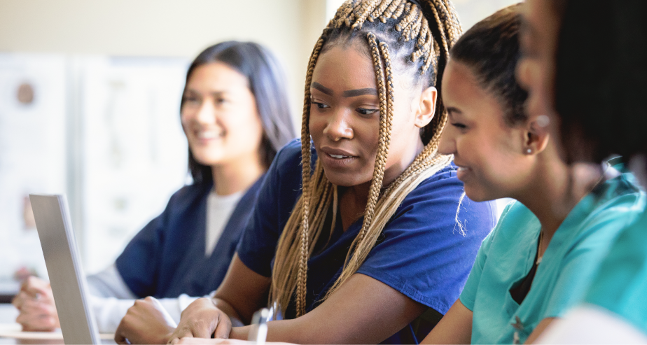 Un grupo diverso de mujeres con bata sentadas alrededor de una mesa, reflejando unidad y colaboración en un entorno médico.
