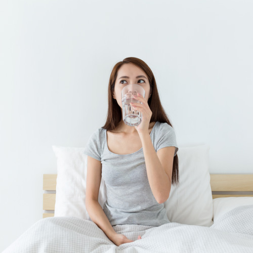 Mujer joven sentada en la cama bebiendo un vaso de agua, inicio saludable del día.