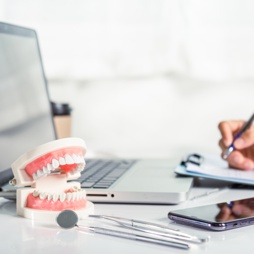 n modelo de mandíbula dental y herramientas de dentista, incluyendo un espejo y un explorador, se encuentran sobre un escritorio. Al fondo, una persona toma notas en un cuaderno, y se puede ver un teléfono móvil y una computadora portátil. La escena sugiere un entorno de estudio o trabajo en el campo de la odontología.